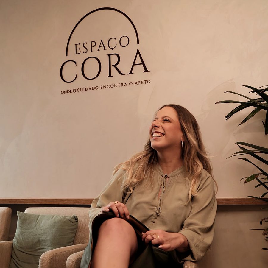 Duda Camargo rindo, sentada numa poltrona, com o logo do Espaço Cora na parede ao fundo