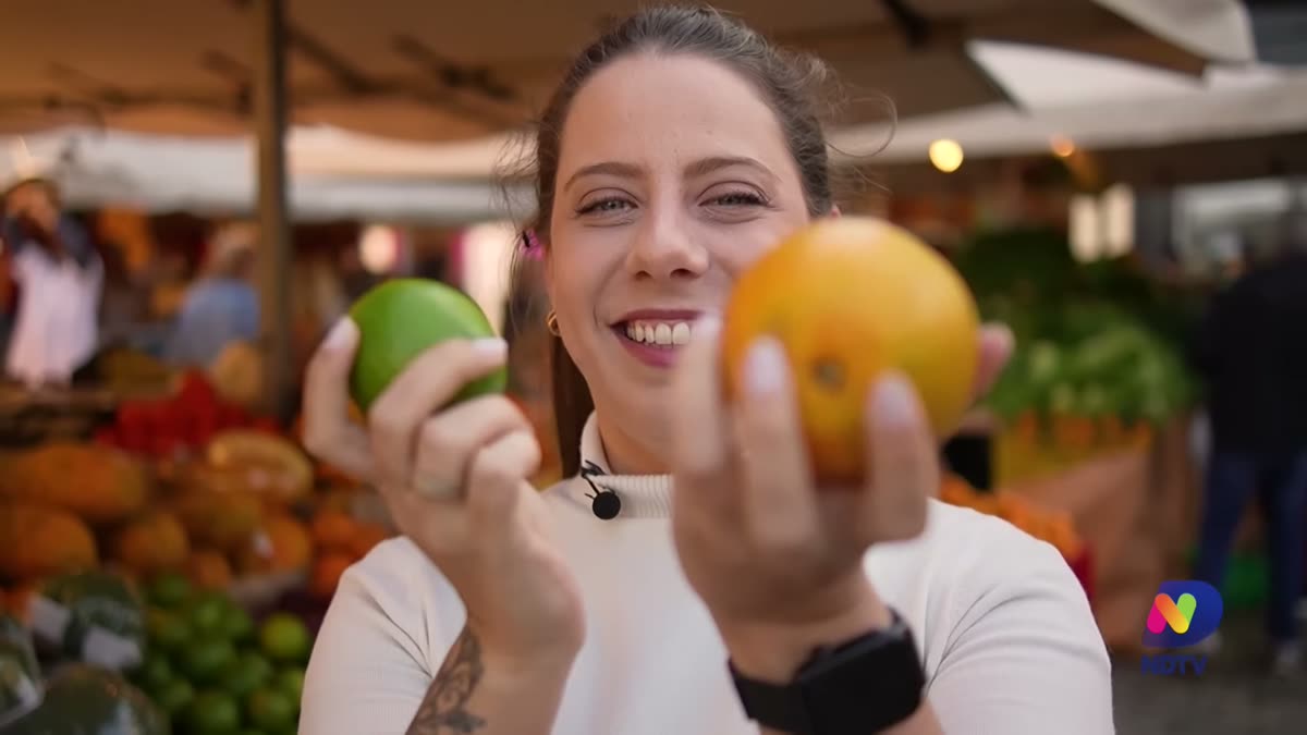 Duda Camargo numa feira, segurando um limão e uma laranja, em reportagem da NDTV